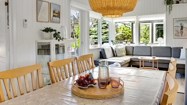 A dining area with a round table, chairs, a wicker pendant light, and a view of the outdoors through large windows.