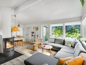 Ein modernes Wohnzimmer mit Kamin, einer Couch, einem Essbereich und großen Fenstern mit Blick auf Grünflächen.