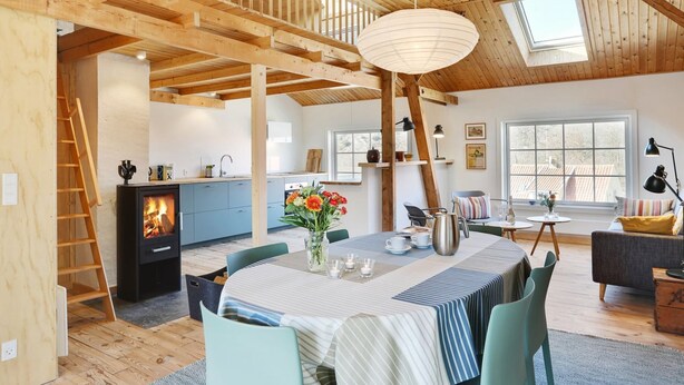 Ein großzügiger Raum mit hohen Decken, Holzbalken, einem Kamin, einem Esstisch mit Stühlen und einer gemütlichen Sitzecke mit Sofa und Kissen.
