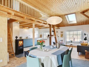Amplia habitación con techo alto, vigas de madera, chimenea, una mesa de comedor con sillas y un rincón de descanso acogedor con sofá y cojines.