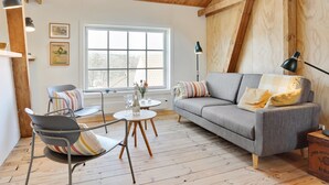 Ein gemütliches Wohnzimmer mit grauem Sofa, einem runden Couchtisch und einem Metallstuhl. Ein großes Fenster lässt viel natürliches Licht herein, und die Decke ist mit Holzbalken versehen.