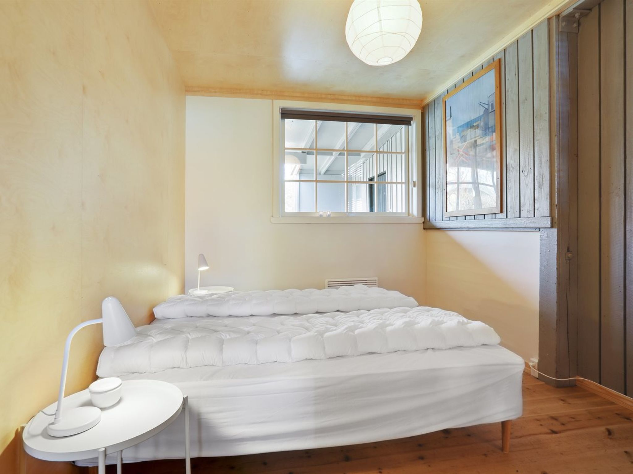 A bedroom with a bed, a bedside table, a lamp, a window, and a view of the outside.