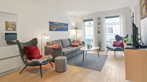 Ett modernt vardagsrum med en grå soffa, två fåtöljer, ett soffbord och en liten pall. På väggen sitter en platt-TV, och det finns även en lampa och konstverk på väggarna.