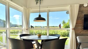Un coin repas comprenant une table ronde, quatre chaises, un luminaire suspendu et une vue sur un champ grâce à de grandes fenêtres.