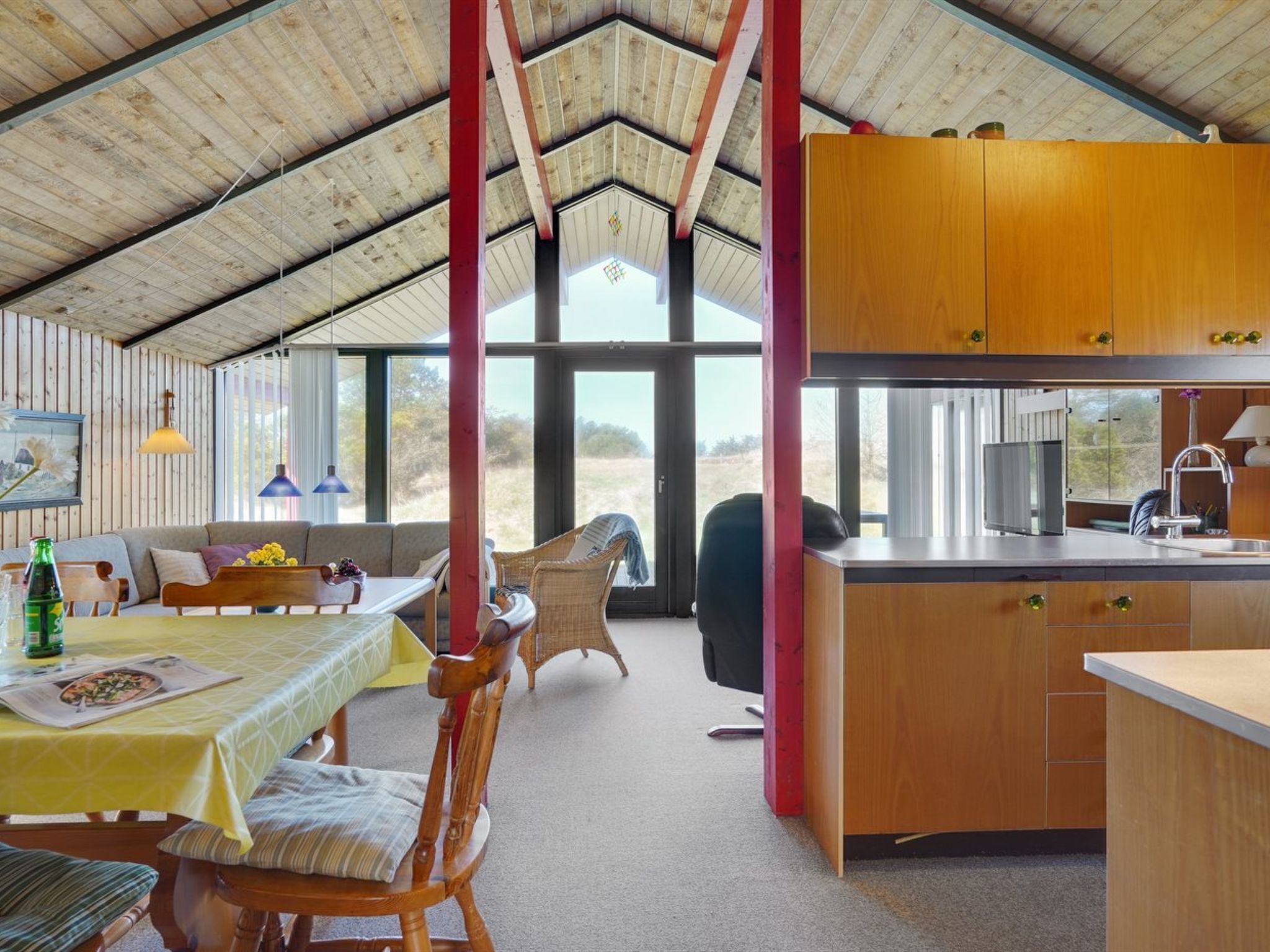 A spacious room with a high ceiling, wooden beams, a dining area with a table and chairs, a kitchen with wooden cabinets, and large windows offering a view of the outdoors.