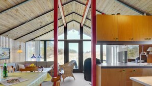 A spacious room with a high ceiling, wooden beams, a dining area with a table and chairs, a kitchen with wooden cabinets, and large windows offering a view of the outdoors. - "Alfrik" - 900m from the sea by Interhome (Blokhus)