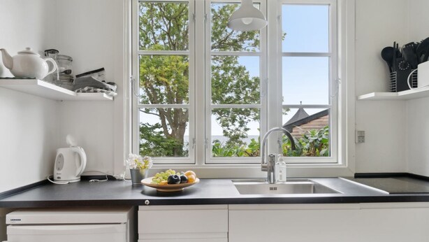 A modern kitchen with a large window, a sink, and a countertop with a kettle and a bowl of fruit. - "Zina" - 25m from the sea by Interhome (Gudhjem)