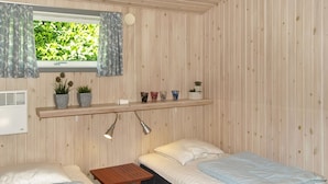 Een kleine houten kamer met twee bedden, een raam met gordijnen, een plank met planten en een kleine tafel.