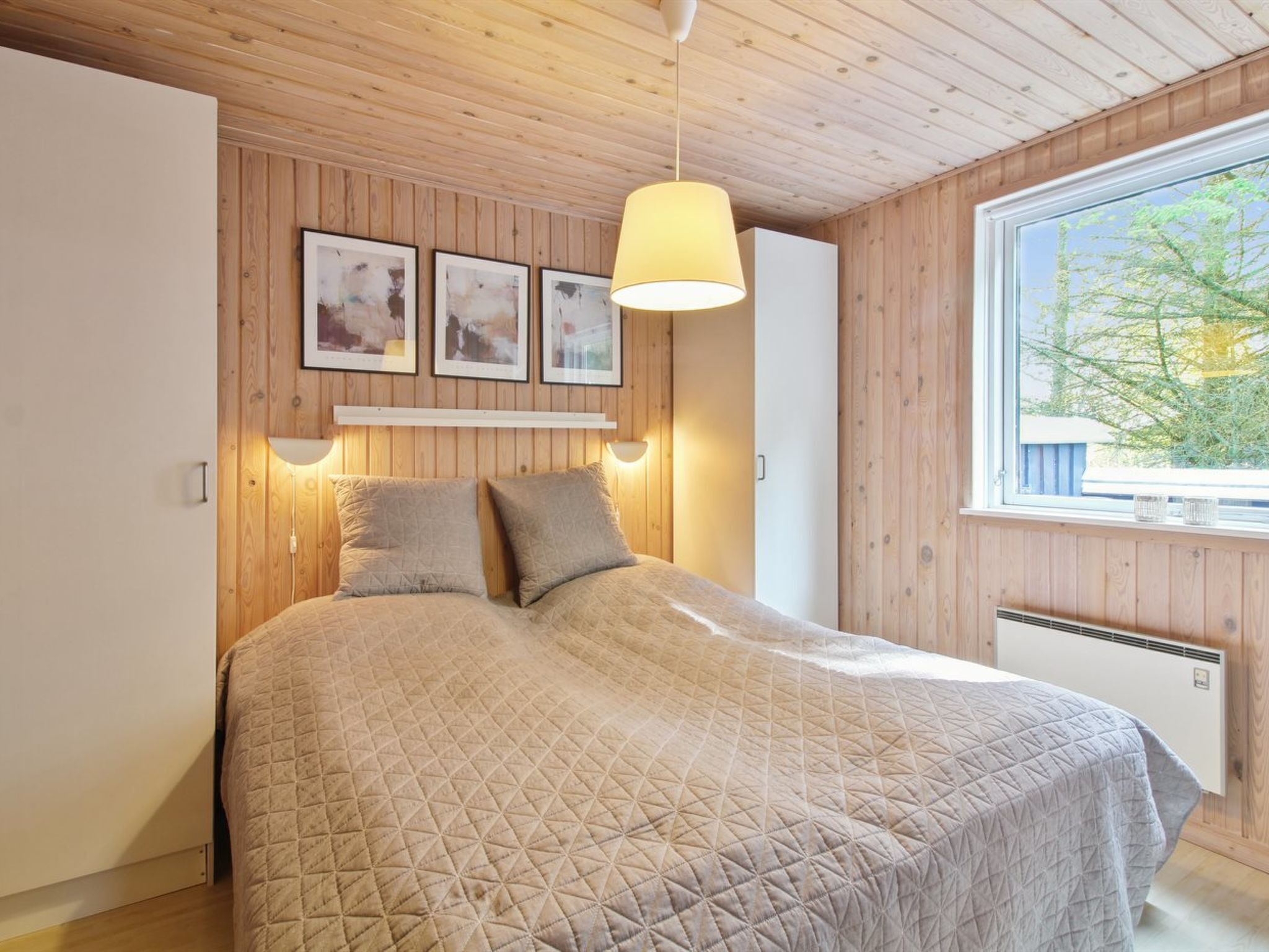 A bedroom with a wooden ceiling, a bed with a quilted cover, a hanging light fixture, and a window with a view of trees.