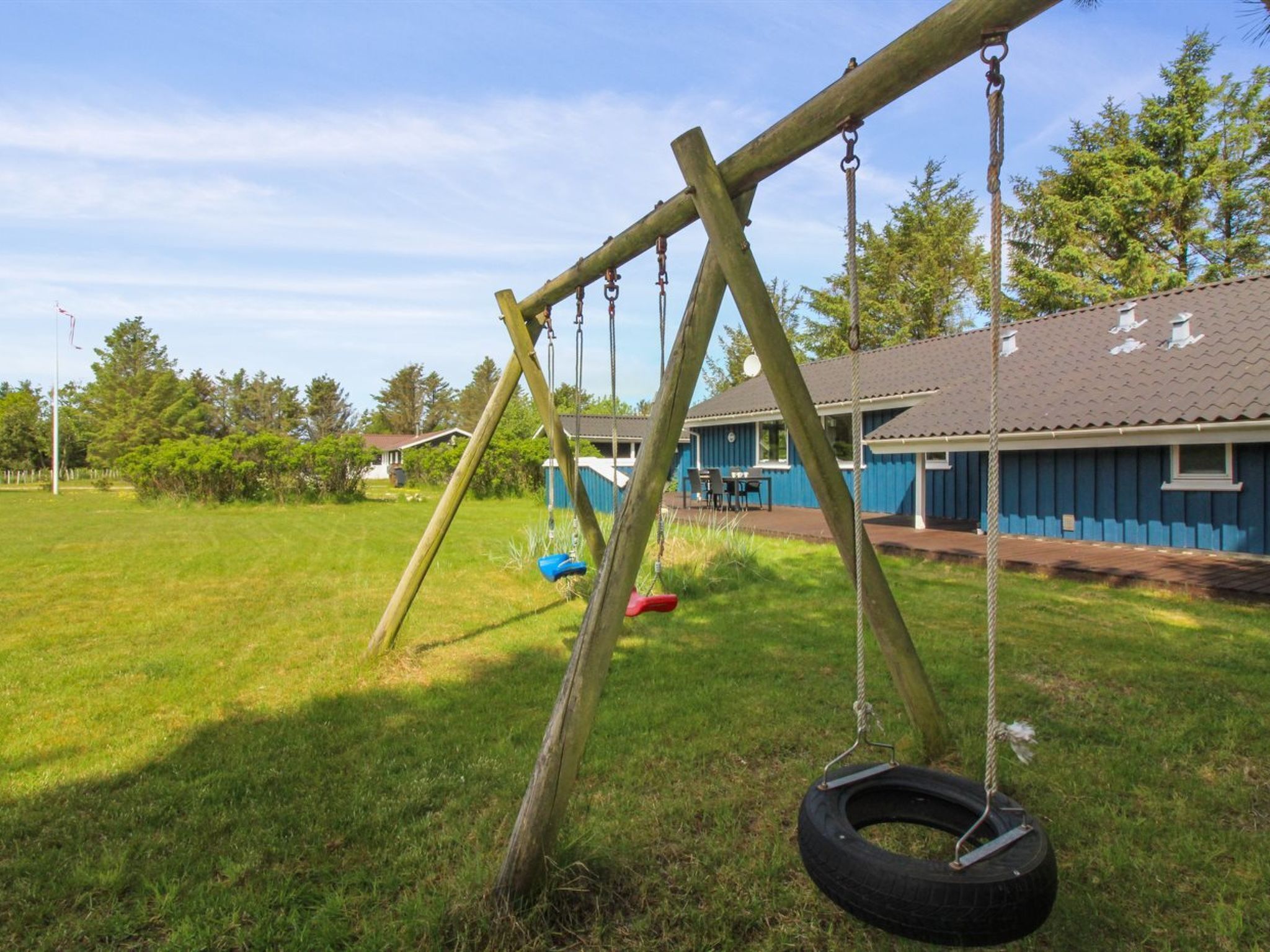 Un ensemble de jeux d’extérieur en bois avec une balançoire faite d’un pneu, un bâtiment bleu avec un toit en tuiles et une cour couverte d’herbe.