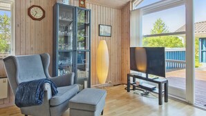 Ett modernt vardagsrum med en grå fåtölj, en pall och ett glasskåp med dekorativa föremål. En platt-TV står på ett TV-bord, och det finns ett stort fönster med utsikt över omgivningen.
