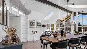 Ein moderner Essbereich mit einem langen Holztisch, schwarzen Stühlen und Pendelleuchten. An der Wand befinden sich gerahmte Bilder und ein großes Fenster, das für natürliches Licht sorgt.