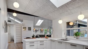 Eine moderne Küche mit weißen Schränken, einer großen Kochinsel, Pendelleuchten und Blick ins Freie durch ein Fenster.