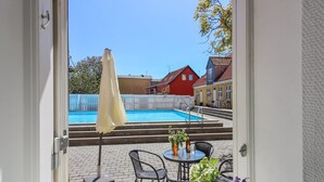 A view of a swimming pool with steps leading to it, surrounded by a white fence. There are houses in the background, and a table with chairs and a bottle on it. - "Thyrne" - 500m from the sea by Interhome (Gudhjem)