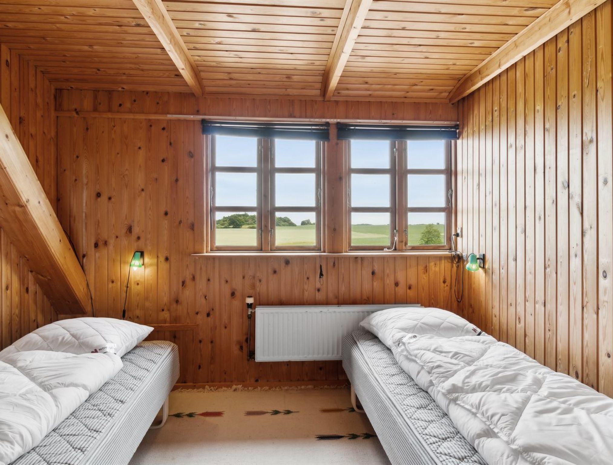 A wooden room with two beds, a radiator, and two windows with views of a green landscape.