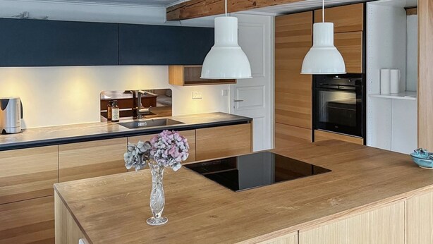 Eine moderne Küche mit Holzschränken, einer zentralen Kochinsel mit schwarischem Induktionskochfeld, Pendelleuchten und einer Vase mit Blumen.