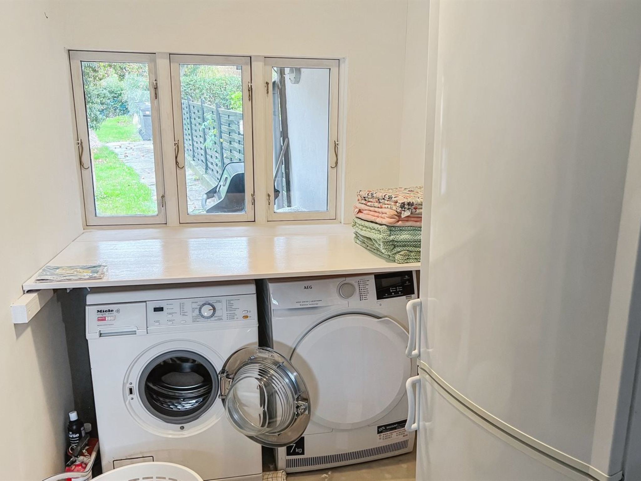A laundry area with two washing machines, a window with a view of a garden, and a stack of folded blankets on the counter.