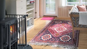 A cozy room with a fireplace, a wooden floor, a patterned rug, and a window with a view of greenery.