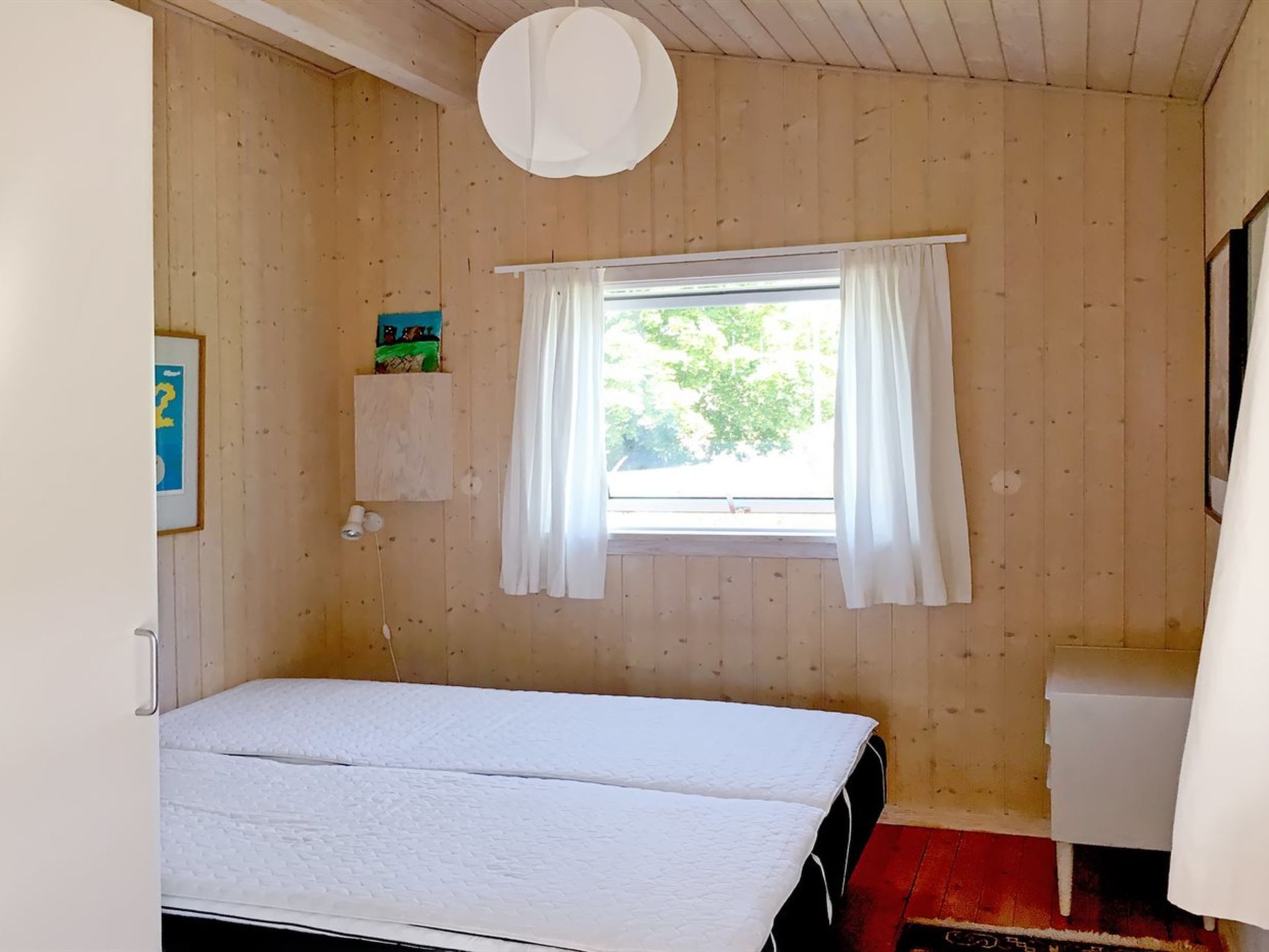 A small, single-bed room with wooden walls, a window with white curtains, a bedside table, and a small shelf with a picture.