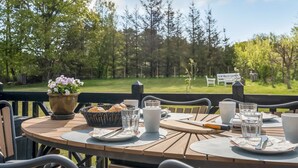 Une table en bois installée à l’extérieur, avec vue sur les arbres, et un panneau visible au fond.