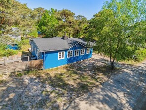 Une maison bleue avec un toit sombre, entourée d’arbres et bordée d’un chemin de terre.