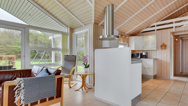 Amplia sala de estar con techo de madera, un ventanal grande, un sofá, una silla, una mesita con un jarrón y una zona de cocina con armarios y microondas.