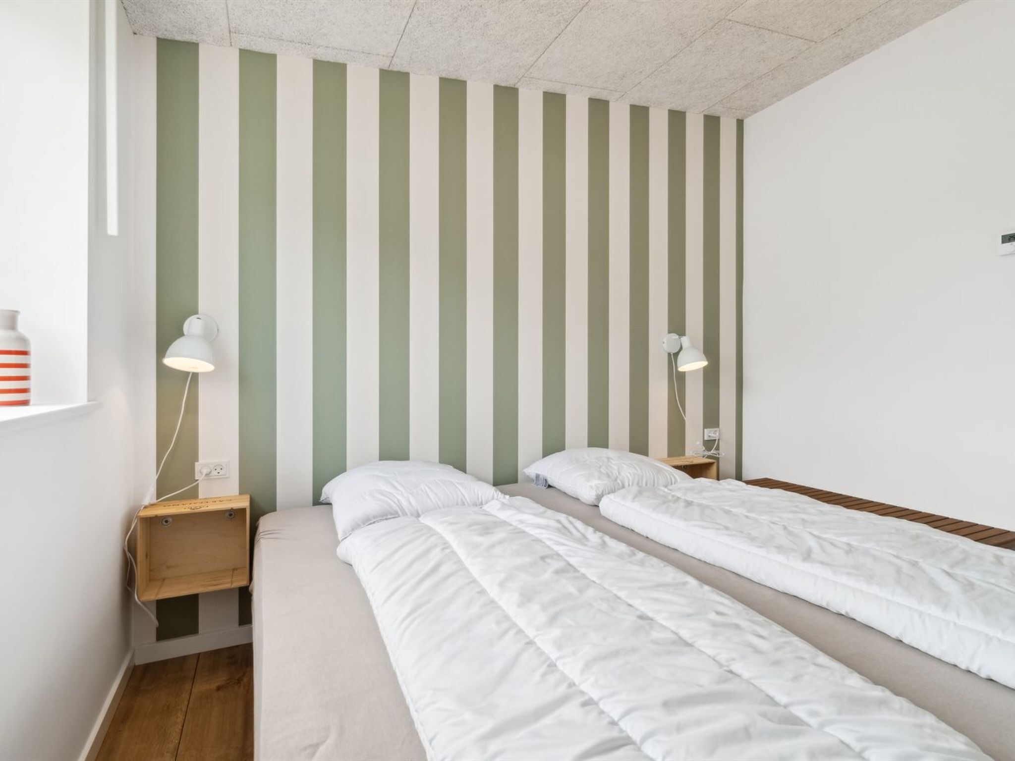 Une chambre avec deux lits simples, une table de chevet en bois, une lampe fixée au mur et une petite étagère avec une bouteille.