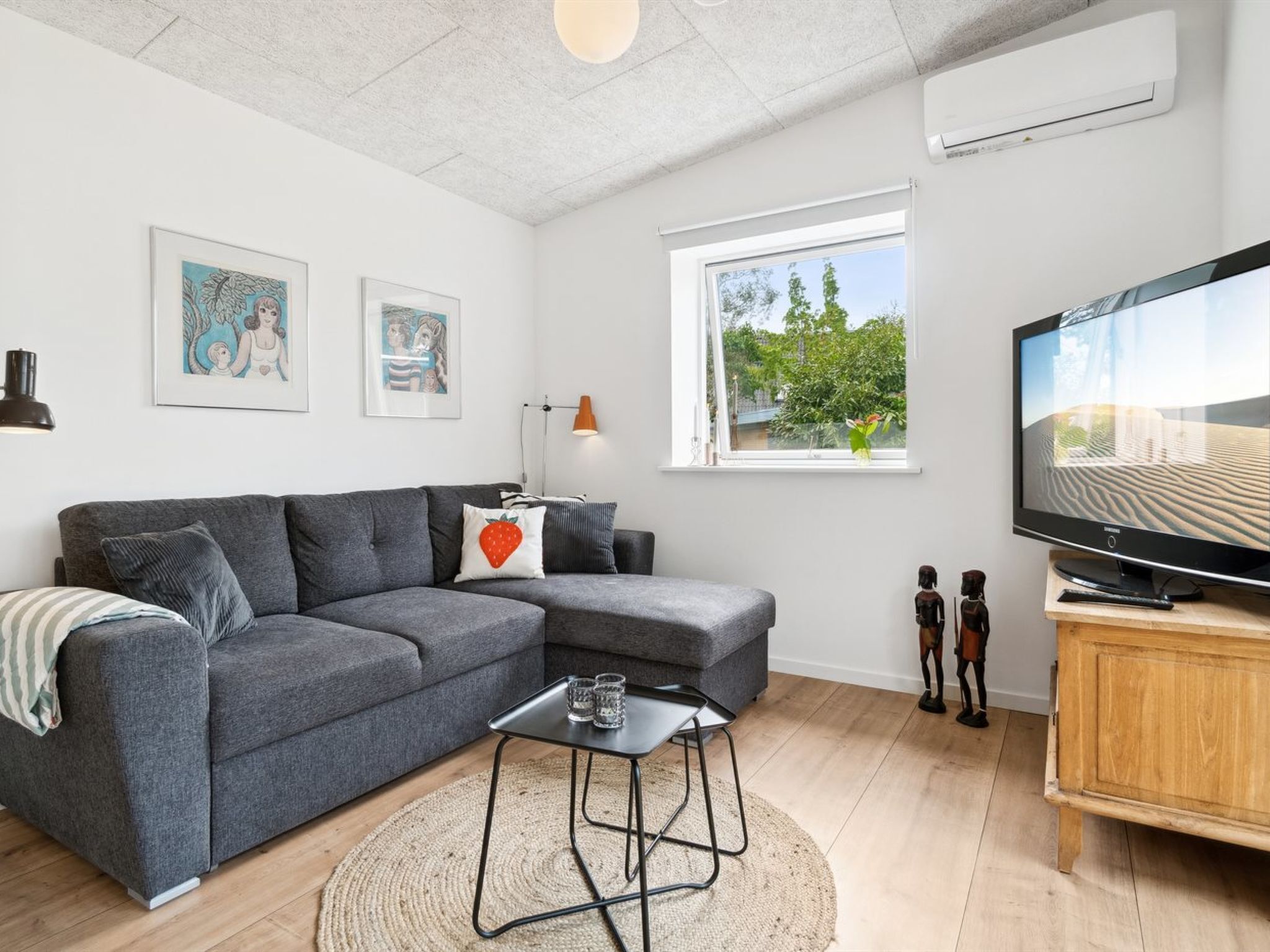 Un salon moderne avec un canapé d’angle gris, une petite table basse, un téléviseur à écran plat et une fenêtre donnant sur un paysage verdoyant.