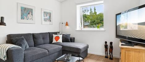 Un salon moderne avec un canapé d’angle gris, une petite table basse, un téléviseur à écran plat et une fenêtre donnant sur un paysage verdoyant.