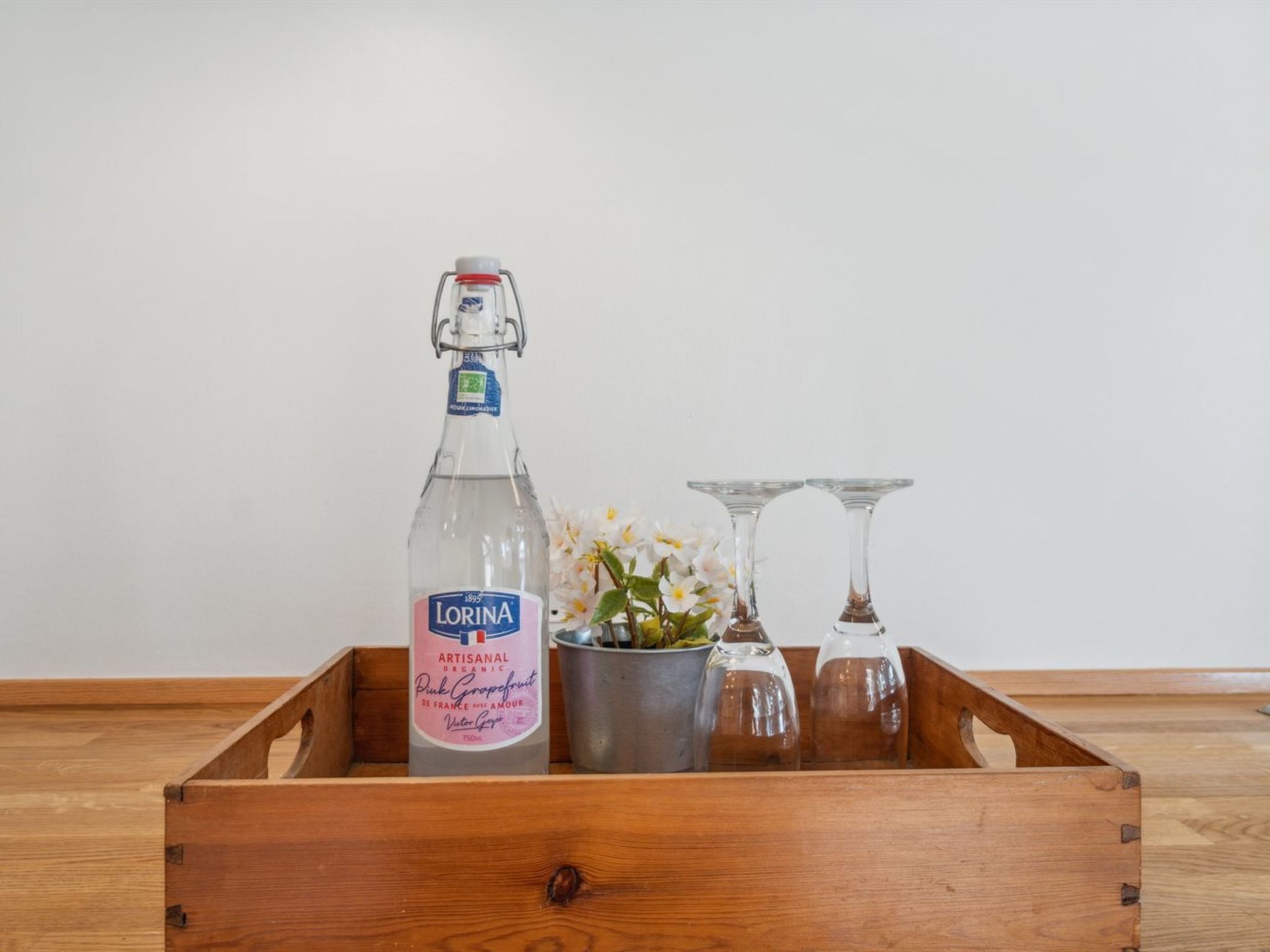 A wooden crate containing a bottle of Lorina Artisanal Pink Grapefruit, a metal bucket, and two glass wine glasses.