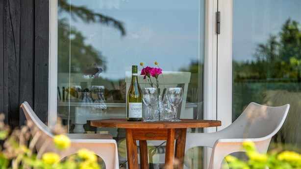 Une table avec une bouteille de vin et des verres, une fleur et une chaise, le tout dans un cadre extérieur ensoleillé.
