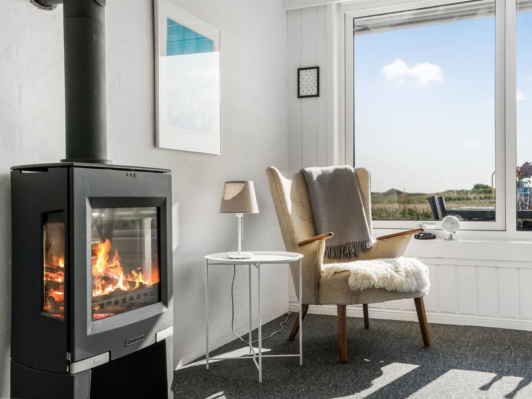 A room with a wood-burning stove, a chair, a small table, and a window with a view.