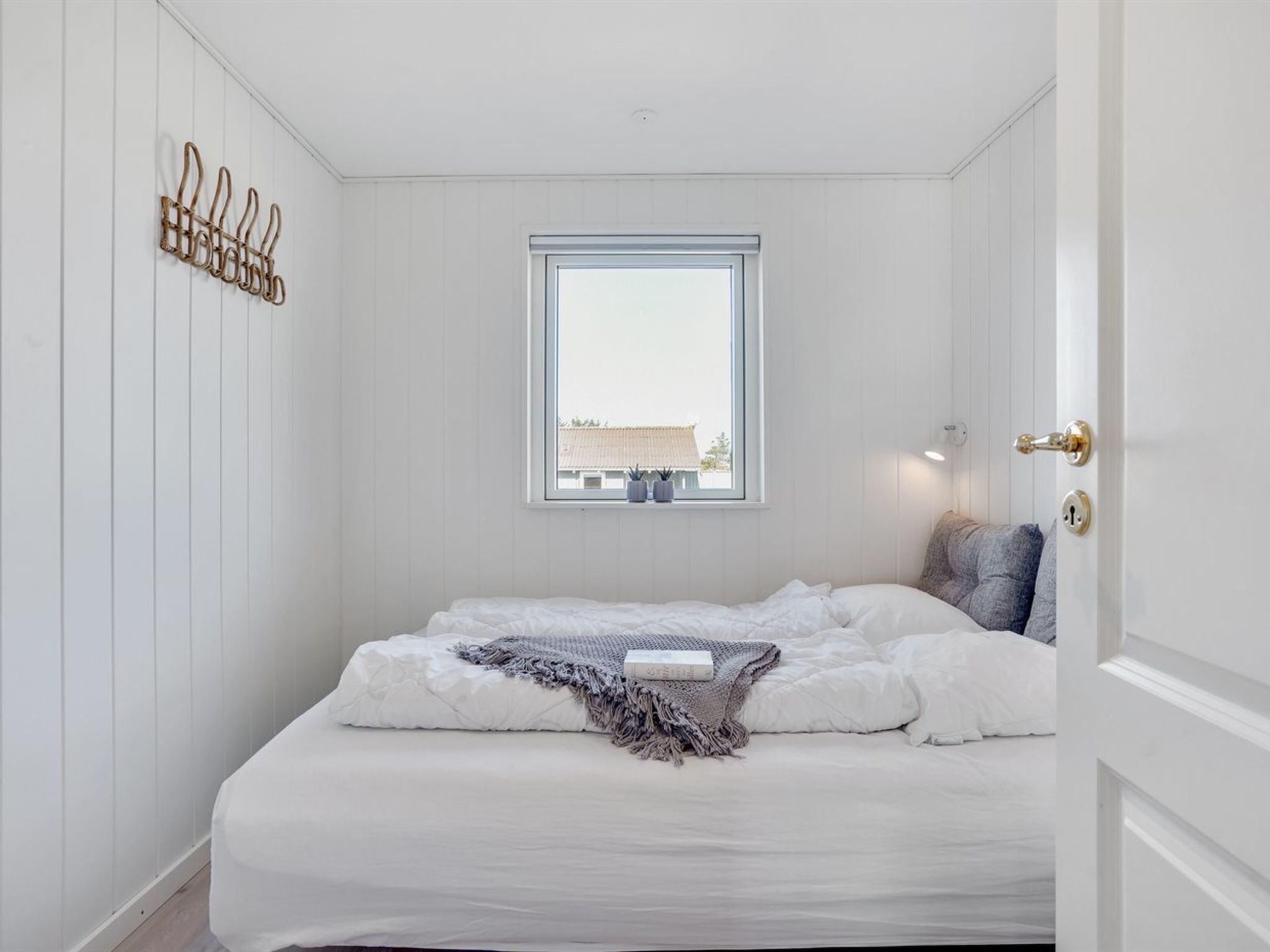 A bedroom with a large window, a bed with white bedding, a gray throw blanket, and a wall-mounted light fixture.