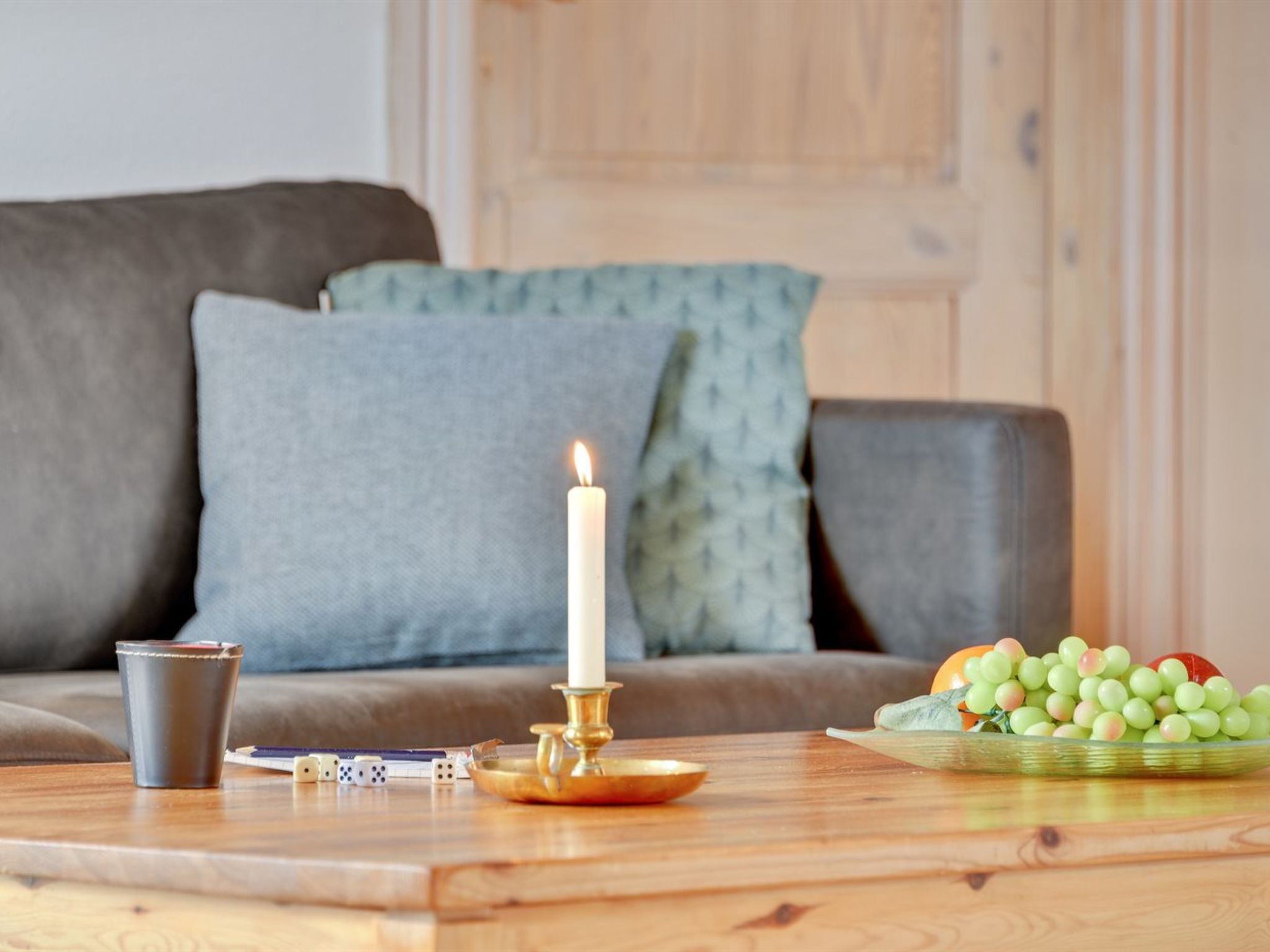 A wooden table with a white candle, dice, and a cup. A bowl of green grapes is on the table.