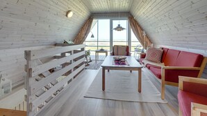 Een knus zolderkamer met een slaande houten schuine kap, een rode bank, een houten tafel en uitzicht op het groen door grote ramen.
