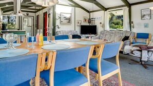 Un vaste espace dédié à la salle à manger, avec une table en bois et des chaises à l’assise bleue, un téléviseur et une grande fenêtre donnant sur un paysage verdoyant.