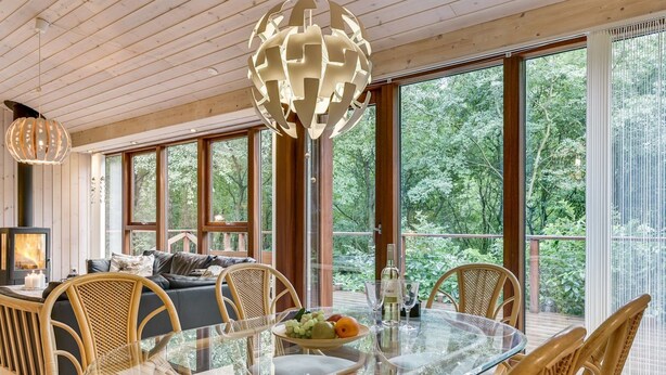 A cozy cabin interior with a glass dining table surrounded by wicker chairs, a fireplace, and large windows offering a view of the forest.