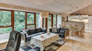 A spacious living area with a large window, a dining table, and a kitchen in the background.