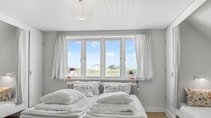 Uma cama de casal com roupa de cama branca, duas mesas de cabeceira com luminárias, uma janela com cortinas e vista para o exterior.
