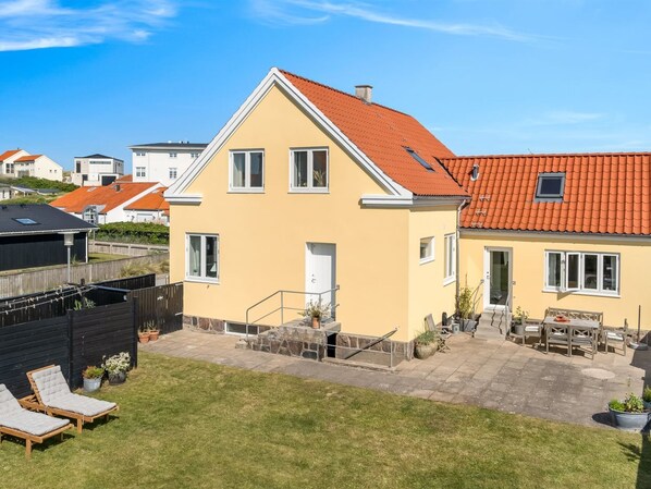 Une maison à deux étages, avec un toit recouvert de tuiles rouges, un petit balcon et un patio aménagé avec du mobilier d’extérieur.