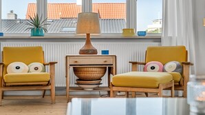 Quarto com duas poltronas amarelas, uma mesa lateral de madeira com abajur, e vista para um edifício com telhados laranja através de grandes janelas.