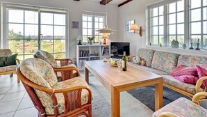 A spacious living room with a wooden table, wicker chairs, a sofa, and large windows offering a view of the outdoors. - "Tordis" - 250m to the fjord by Interhome (Roslev)