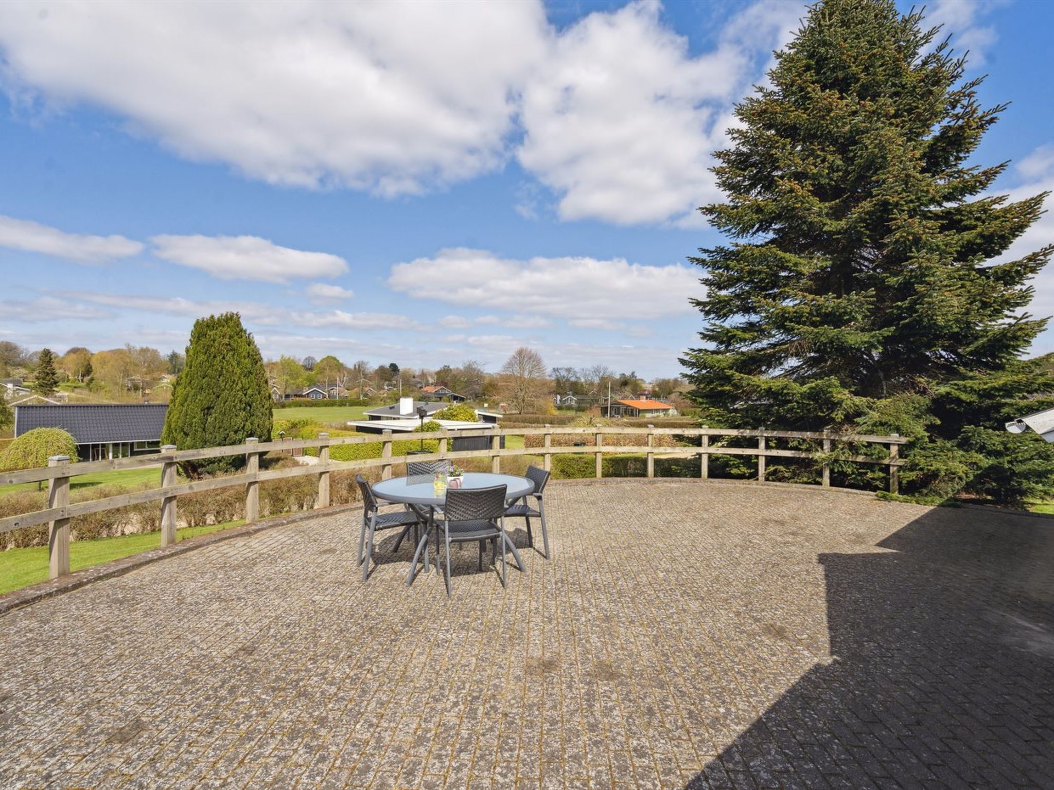 Eine Terrasse mit Tisch und Stühlen, umgeben von einem Holzzaun, mit Blick auf eine Wohngegend mit Häusern und einem großen Baum.