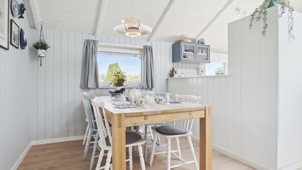Un coin repas avec une table en bois et des chaises blanches, un luminaire suspendu et une fenêtre avec des rideaux.