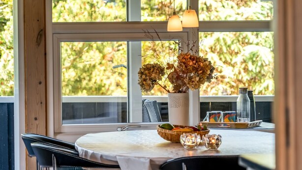 Un coin repas avec une table dressée pour deux personnes, un vase contenant des fleurs séchées et une vue sur les arbres à travers de grandes fenêtres.
