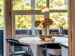 A dining area with a table set for two, a vase with dried flowers, and a view of trees through large windows.
