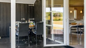 Une porte-fenêtre coulissante donne sur un espace repas comprenant une table et des chaises, et offre une vue sur l’extérieur. Le mur extérieur est en bois.