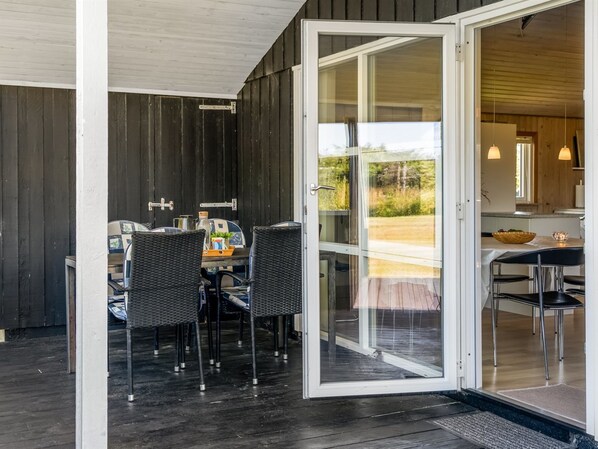 An open sliding glass door leading to a dining area with a table and chairs, a view of the outdoors, and a wooden exterior wall.