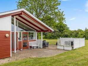 Ein kleines, eingeschossiges Gebäude mit rotem und weißem Anstrich, einer überdachten Terrasse mit Bank und Grill sowie einem weißen Zaun.