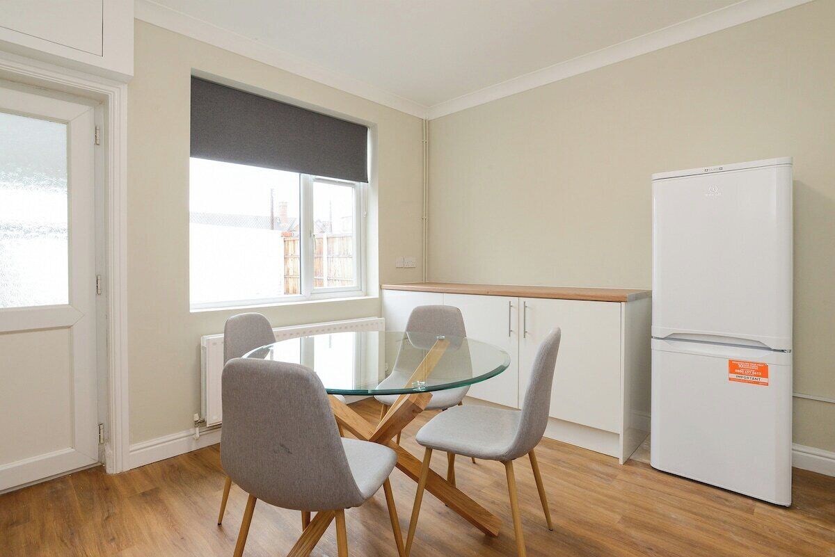 Une cuisine moderne avec une table à manger en verre, des chaises grises, un réfrigérateur blanc et une fenêtre dotée d’un store noir.